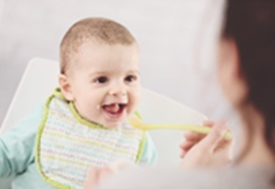 Person spoon feeding a baby. Baby is happy and making eye contact with the caregiver. Their mouth is open as the spoon is offered.